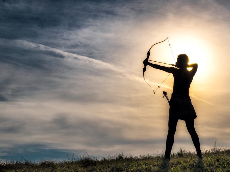 Photo of person in silhouette shooting an arrow into the sky at sunrise.
