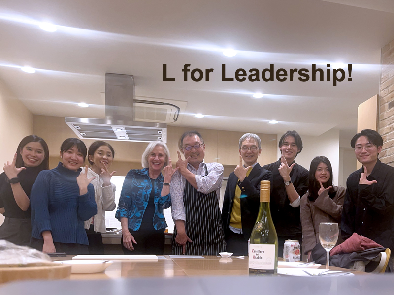 Cherrey and Higano standing with others behind a counter. All making an "L" with their fingers. L for Leadership!