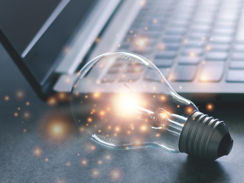 Photo of a light bulb with sparkles coming out of it laying on a desk next to a lap top computer